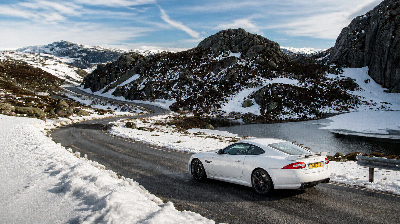 Carro branco circula por estrada sinuosa rodeada de montanhas e neve num cenário de inverno.