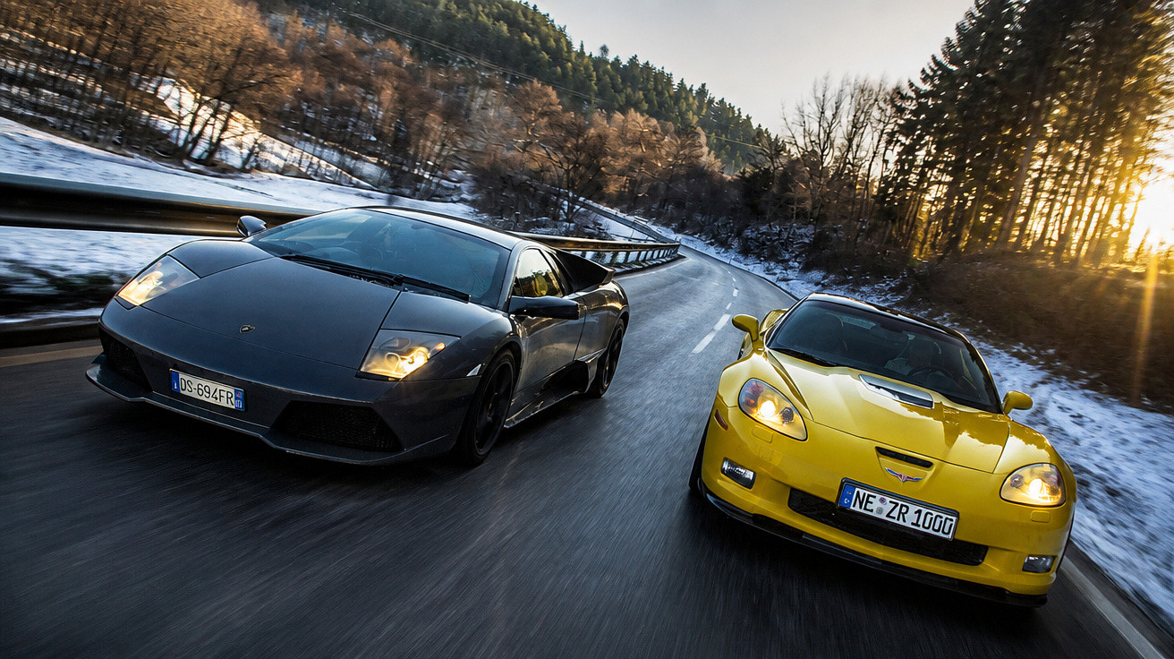 Dois carros desportivos, um preto e um amarelo, a circular numa estrada curva entre árvores e neve.