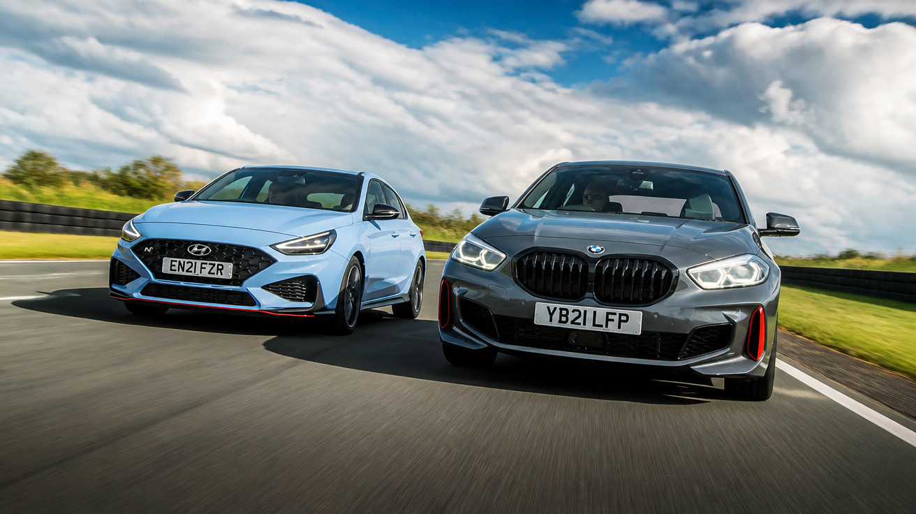 Dois carros desportivos a circular em pista, um cinzento BMW e outro azul Hyundai, sob céu nublado.